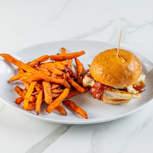 Chicken sandwich with bacon and slaw, served with a side of sweet potato fries on a white plate.