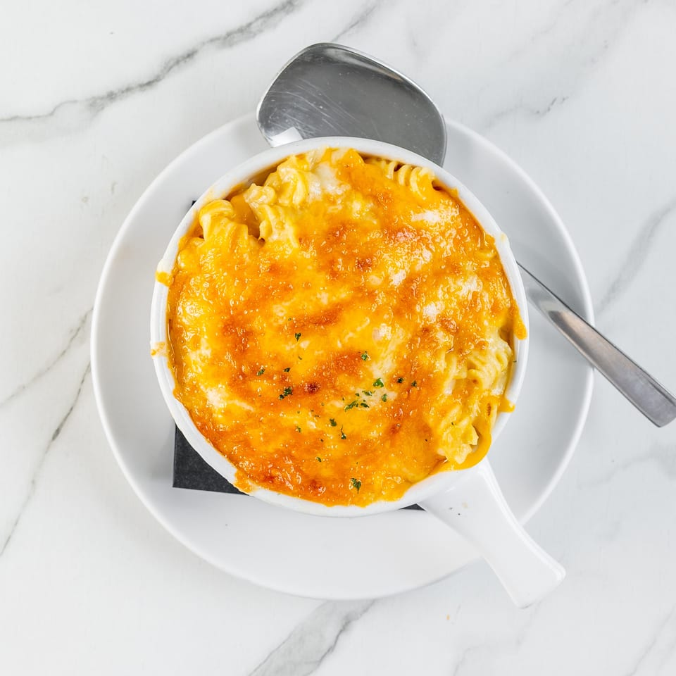 Baked macaroni and cheese topped with golden melted cheese and herbs, served in a white dish with spoon on marble.