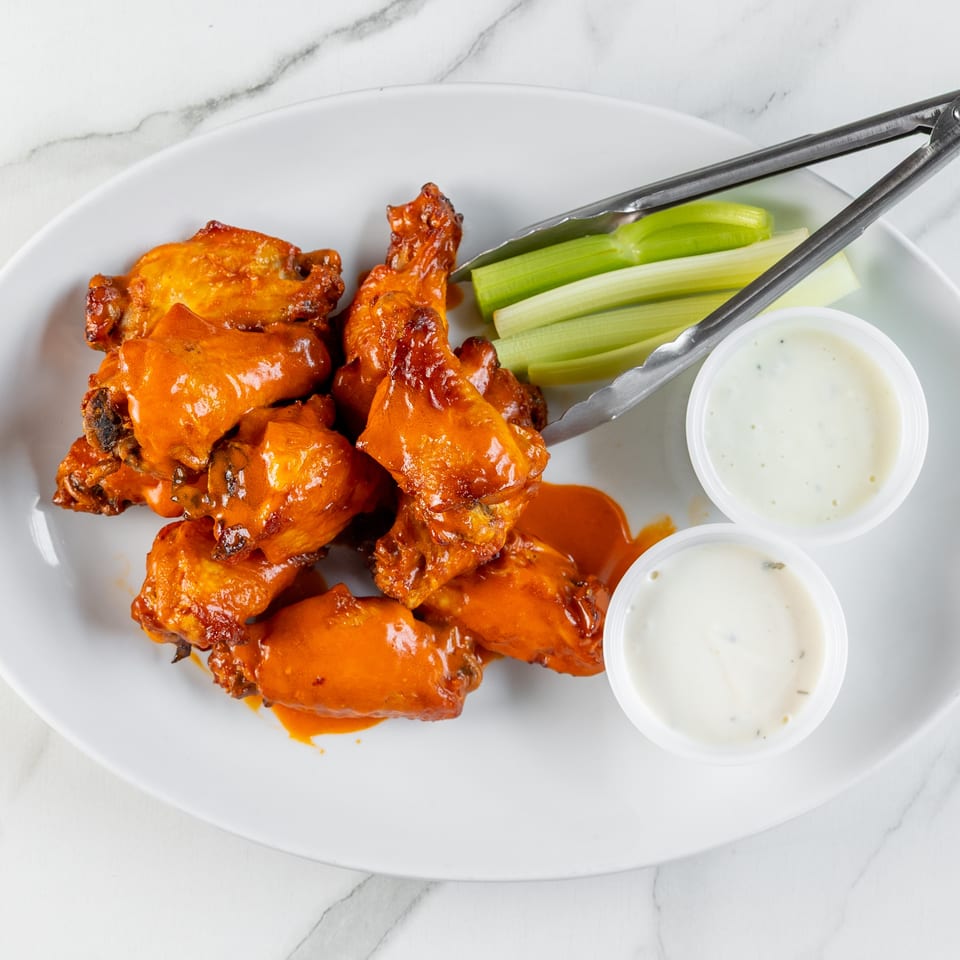 Plate of saucy buffalo wings, celery sticks, two dipping sauces, and tongs on a white marble surface.