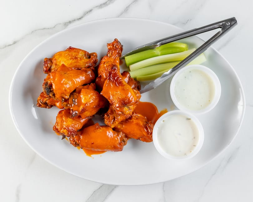 Plate of saucy buffalo wings, celery sticks, two dipping sauces, and tongs on a white marble surface.
