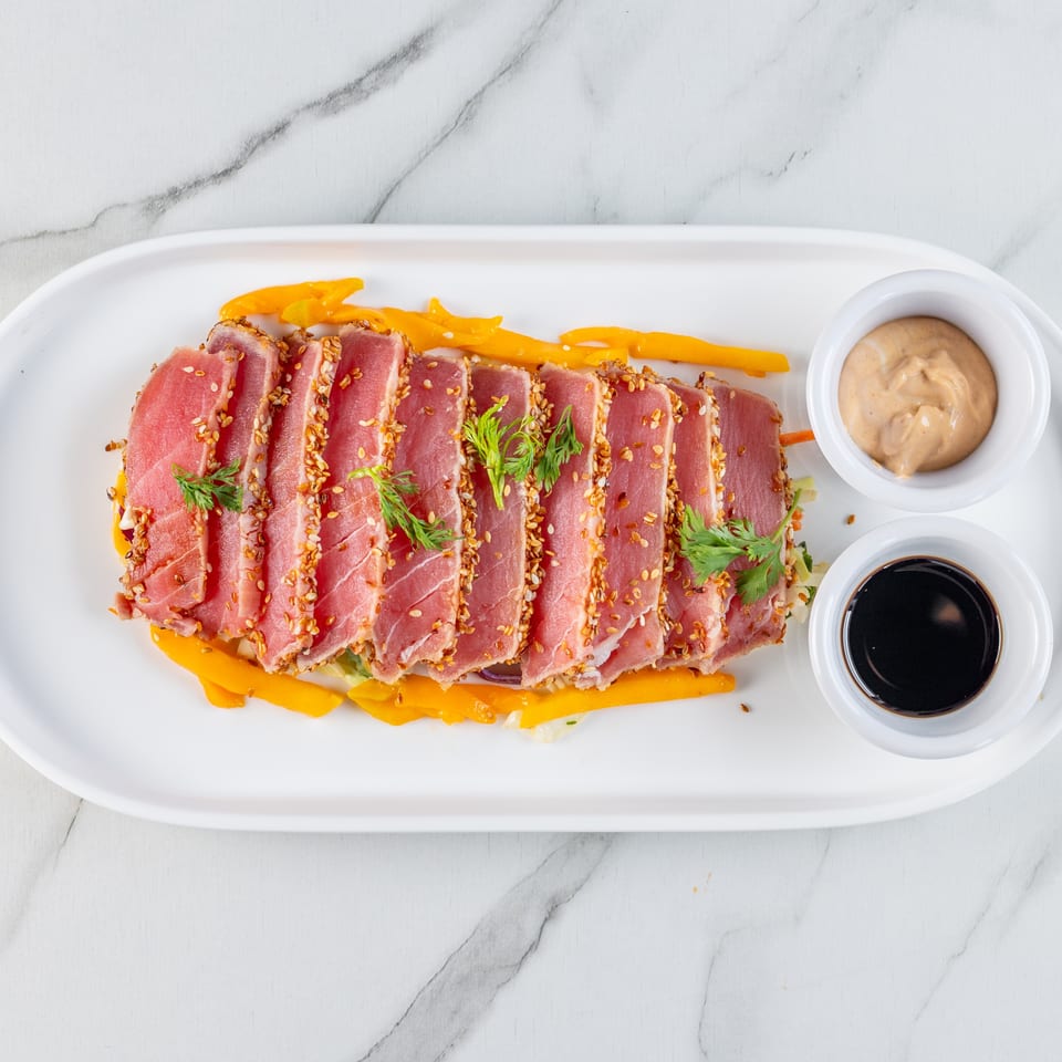 Seared tuna slices with sesame seeds, yellow strips, greens, and two dipping sauces on a white plate.