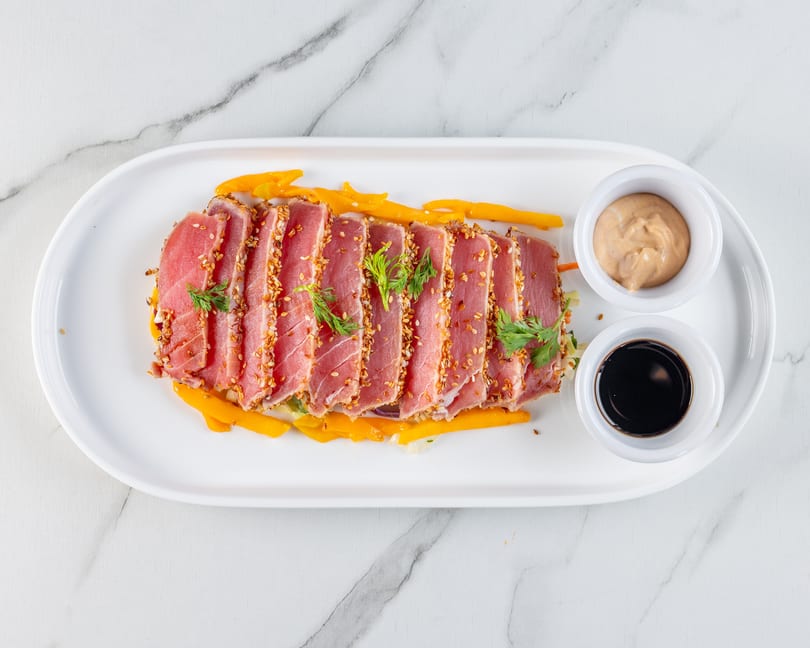 Seared tuna slices with sesame seeds, yellow strips, greens, and two dipping sauces on a white plate.