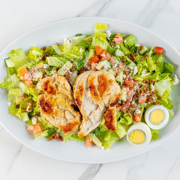 Grilled chicken salad with crisp lettuce, tomatoes, cucumbers, bacon, crumbled cheese, and hard-boiled eggs.