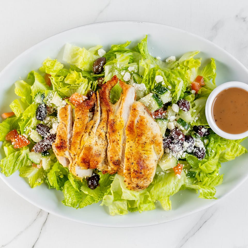 Grilled chicken salad with lettuce, olives, cucumbers, tomatoes, feta, and dressing.