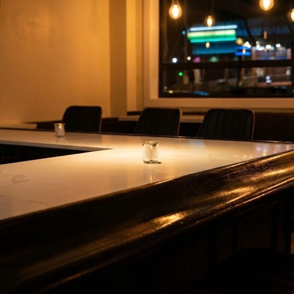 A dimly lit bar area with a white marble countertop, dark chairs, and a street view outside.